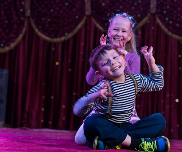 Two children sit on stage making signs with their hands at a theatre